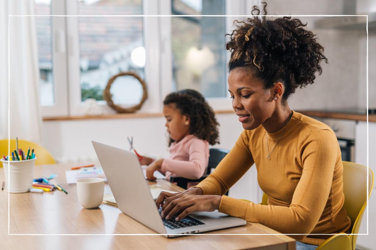  Mum working on laptop at home with daughter sitting beside her at the table. 