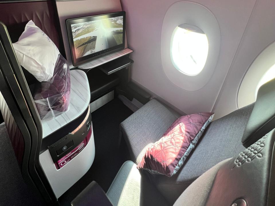 One business class seat on a Qatar Airbus A350, with a burgundy cushion, a window view, and a live feed of the plane on the display screen.