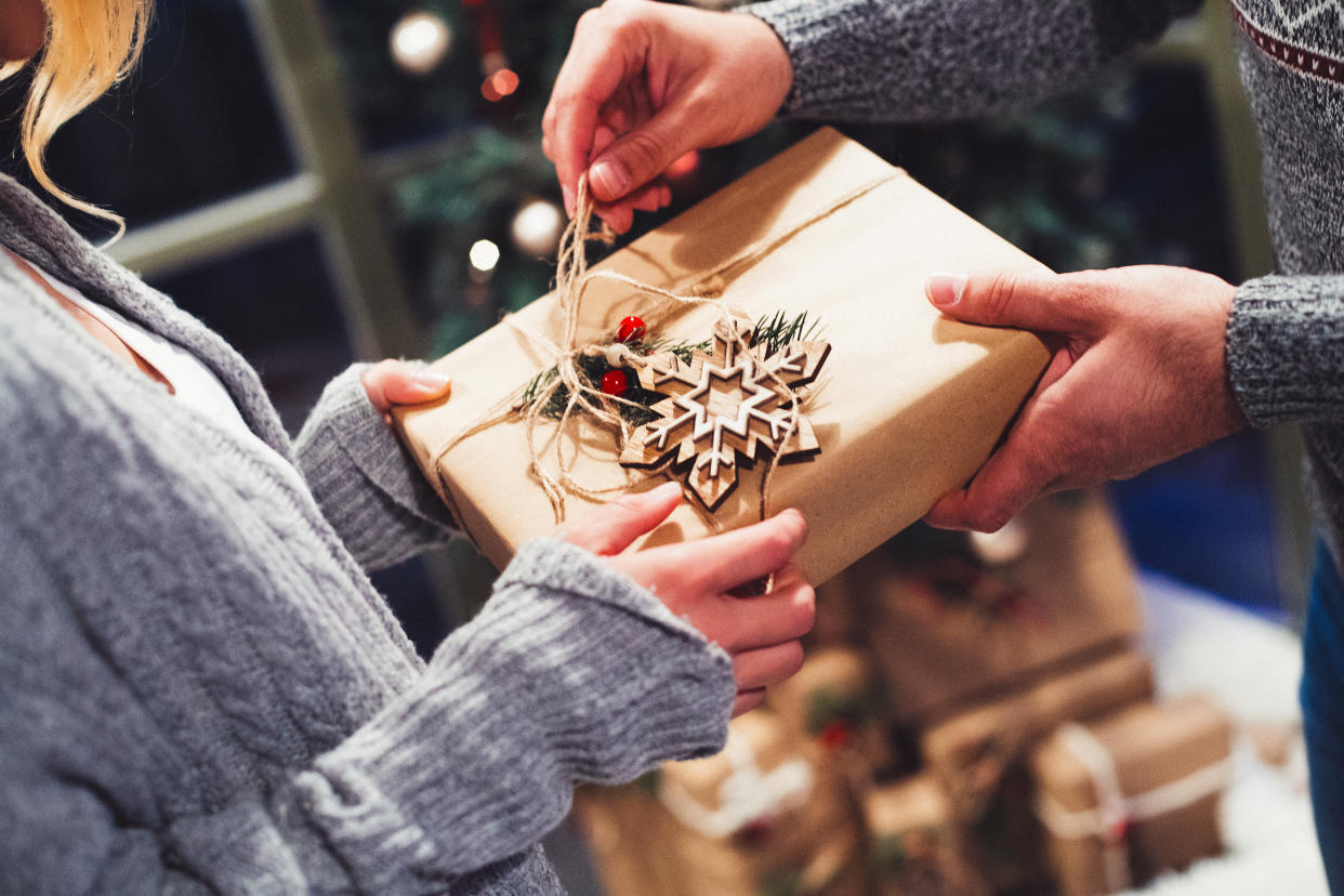 A close-up shot of a man receiving a Christmas gift from a woman. The present is wrapped in kraft paper, decorated with a wooden snowflake and a peace of a pine tree branch and tied with manila rope. Using natural and recyclable materials is one of the ways to make your Christmas environmentally friendly.