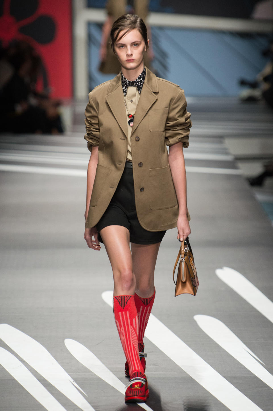 <p><i>Model wears a tan blazer, black shorts, and red knee-high socks from the SS18 Prada collection. (Photo: ImaxTree) </i></p>