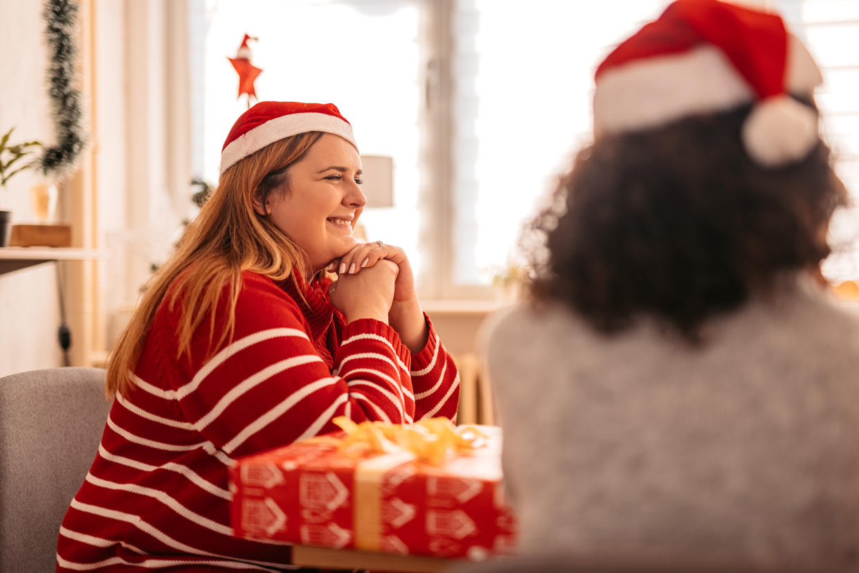 A smiling plus-size woman wearing a Santa hat sits on a couch next to a stack of wrapped presents, with another woman in a Santa hat nearby.