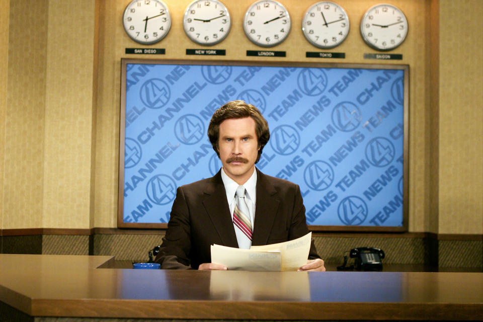 Will Ferrell sits with papers behind a news desk