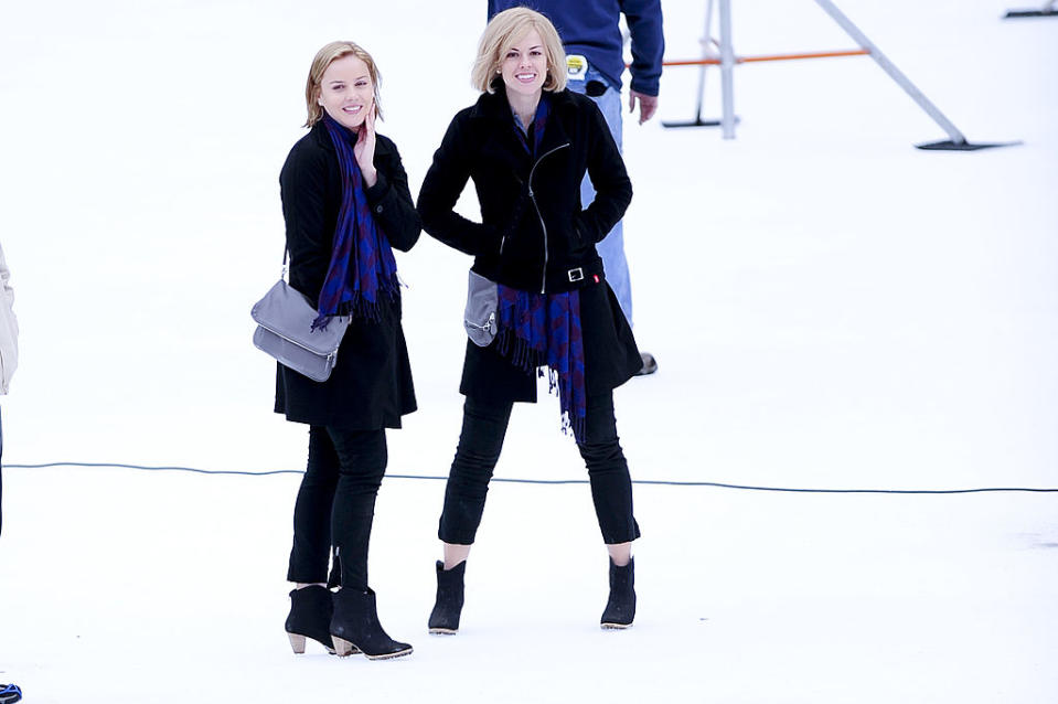 Two women in black coats and boots smiling, standing on snow with blue scarves