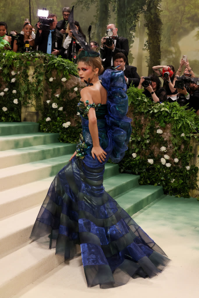 Zendaya in a layered gown posing on steps at an event with photographers in background