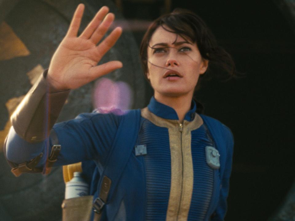 ella purnell as lucy in fallout, pushing aside a panel and wearing a blue and yellow dirty jumpsuit