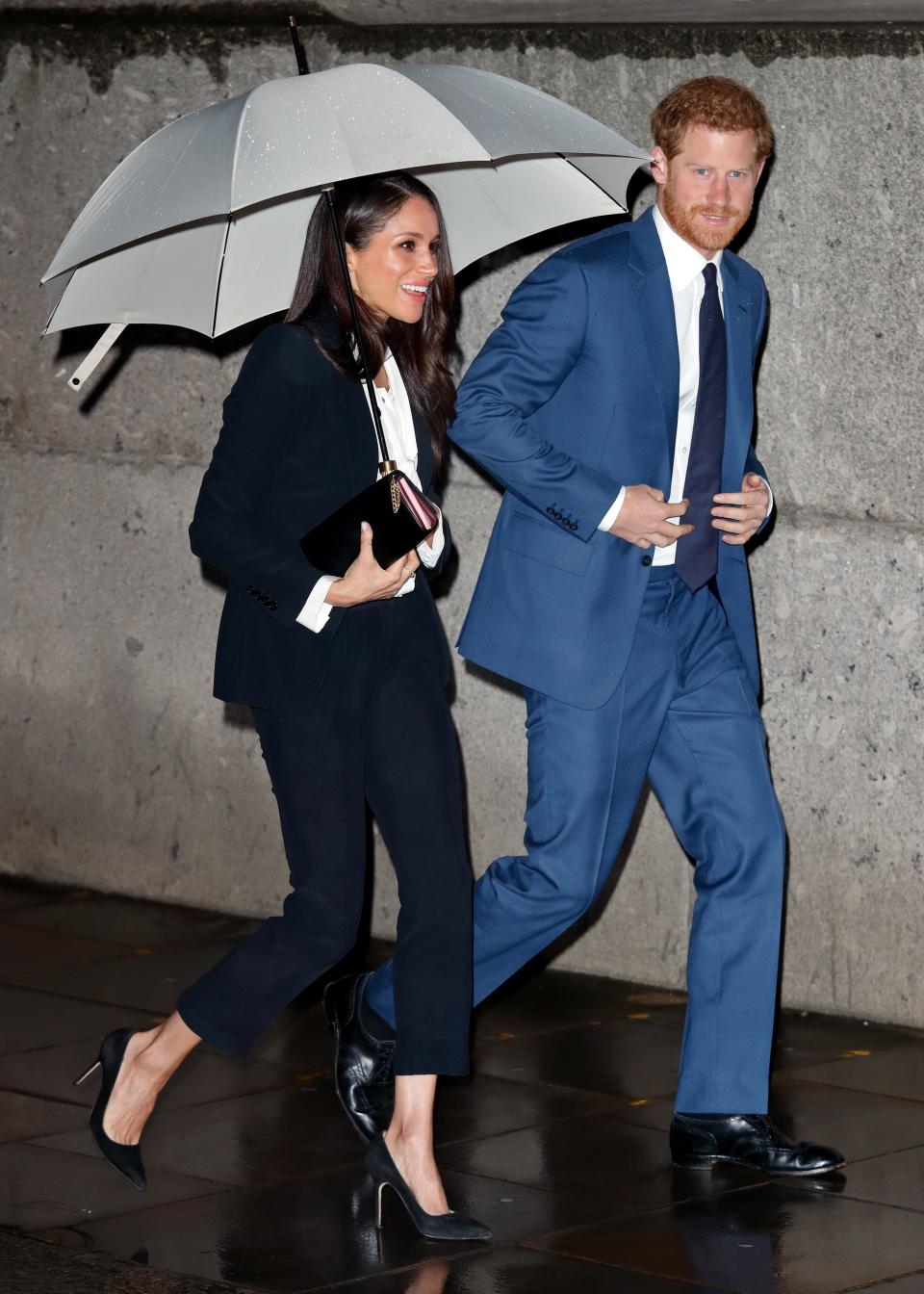 Meghan in a dark slim suit and heels holding an umbrella and Harry in a light blue suit and darker blue tie.