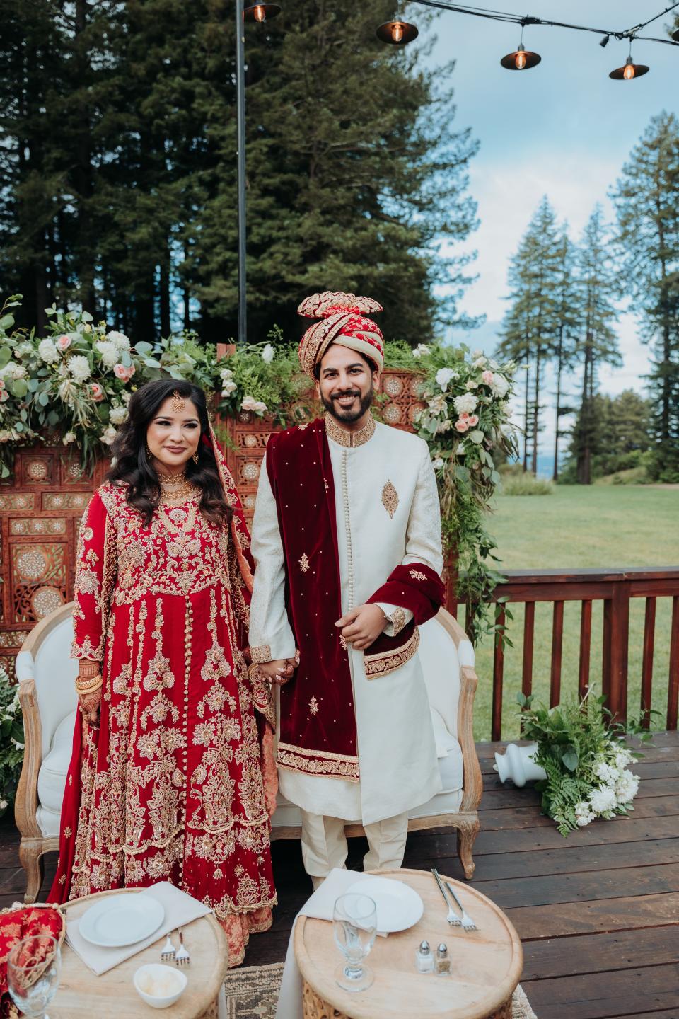 Bride and groom dressed in red and white standing in front of flowers (Uzma Saeed)