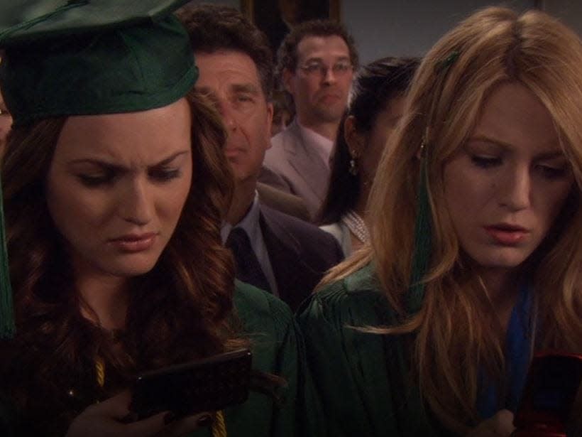 blair and serena looking down at their phones during their high school graduation ceremony