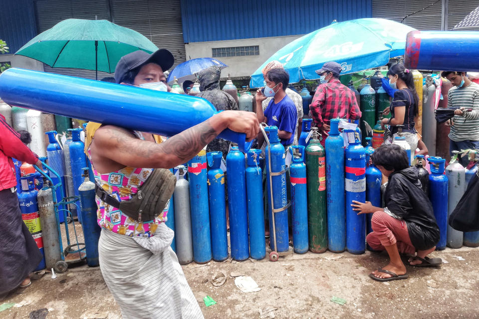 FILE - In this July 28, 2021, file photo, A man carries an oxygen tank while walking past people waiting with oxygen tanks in need of refill outside the Naing oxygen factory at the South Dagon industrial zone in Yangon, Myanmar. Supplies of medical oxygen are running low, and the government has put restrictions on its private sale in many places, saying it is trying to prevent hoarding. (AP Photo, File)