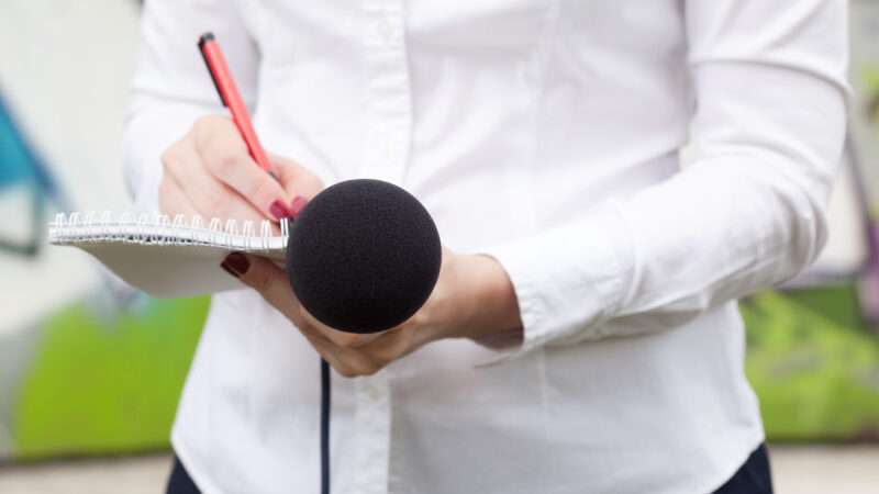 A reporter takes notes with a pad and pen while holding a microphone.