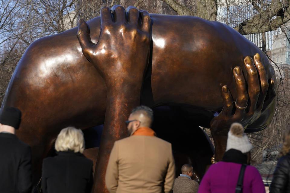 People stand near the 20-foot-high bronze sculpture "The Embrace," a memorial to Dr. Martin Luther King Jr. and Coretta Scott King, in the Boston Common, Tuesday, Jan. 10, 2023, in Boston. The sculpture, consisting of four intertwined arms, was inspired by a photo of the Kings embracing when MLK learned he had won the Nobel Peace Prize in 1964. The statue is to be unveiled during ceremonies Friday, Jan. 13, 2023. (AP Photo/Steven Senne)