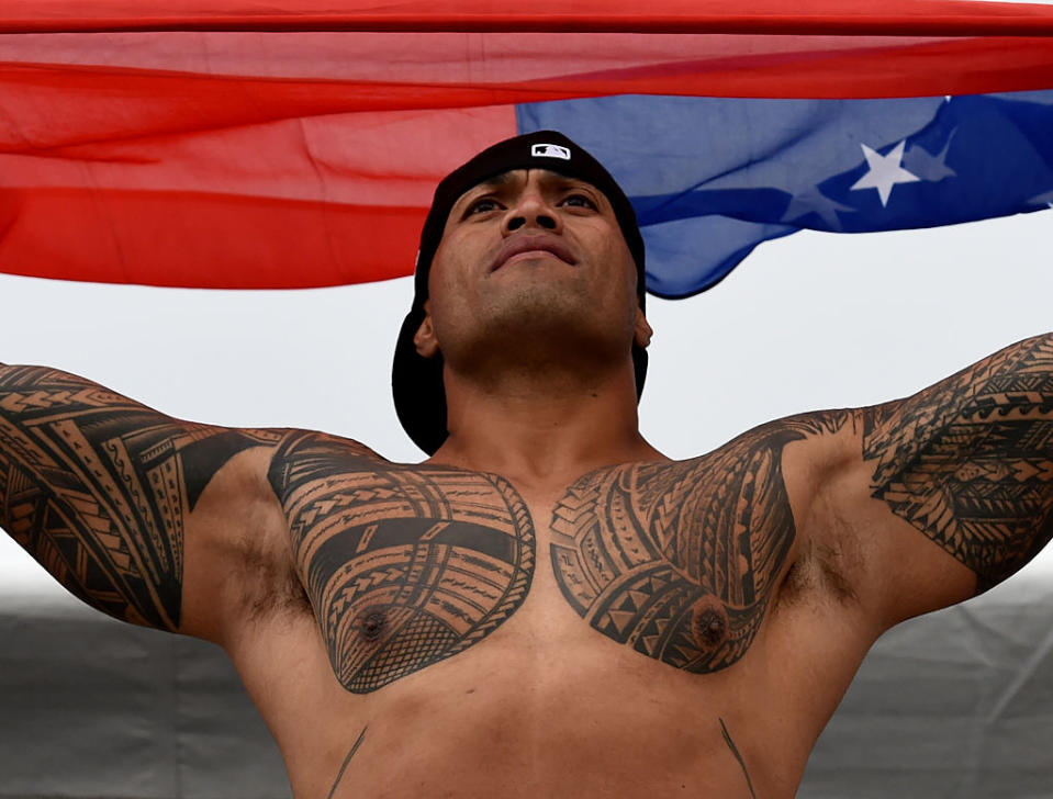 Person with traditional tattoos raises arms under a Samoan flag