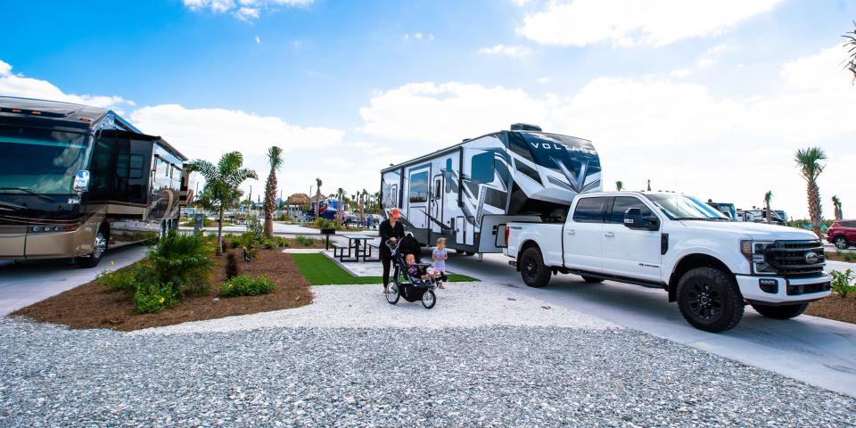 A travel trailer towed by a pickup truck parked at its RV site. There's a person pushing a stroller walking next to a young child next to the RV.