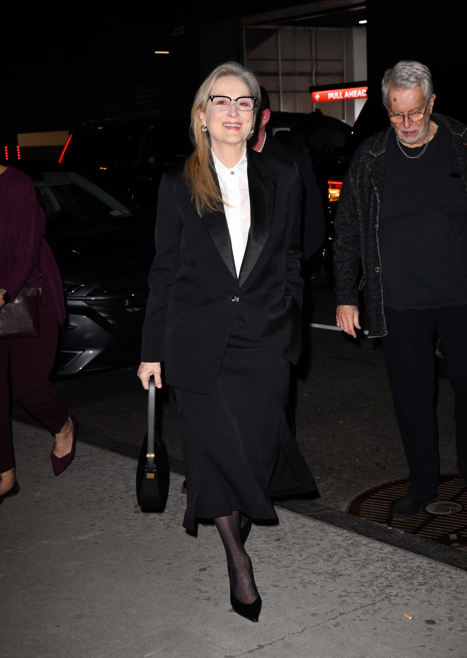 Woman in a black suit with a white shirt, glasses, carrying a purse, walking with a man in a gray sweater