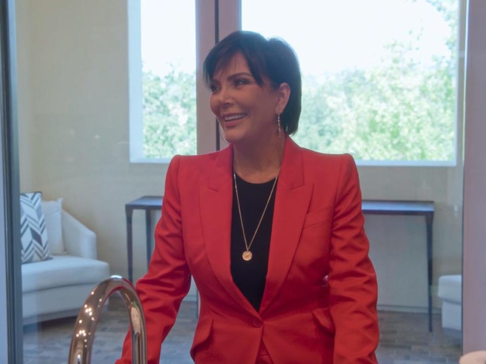 kris jenner smiling while wearing a pink blazer over a black shirt, standing in front of a sun room with a sunny window behind her