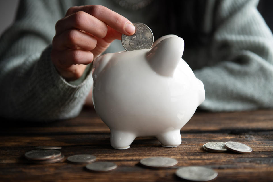 Hand placing a coin into a white piggy bank, with more coins scattered around on a wooden surface