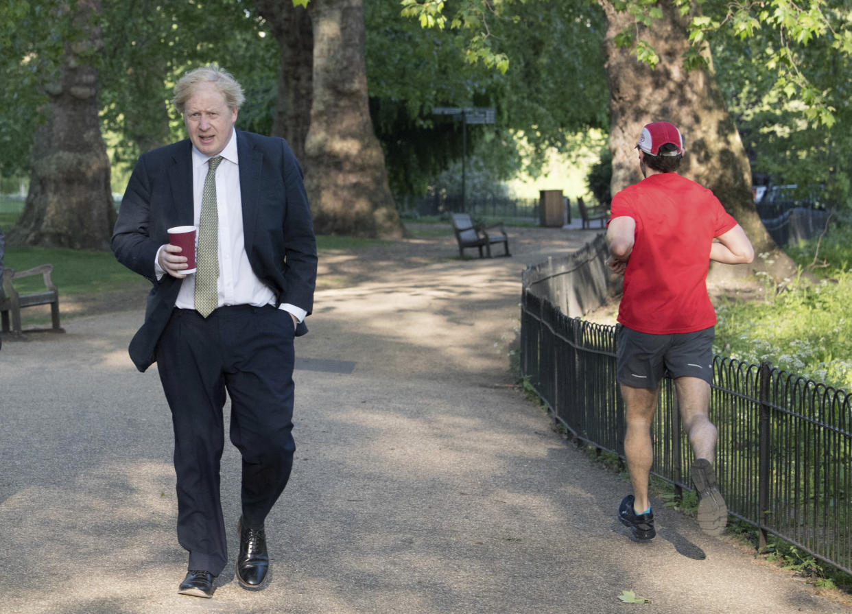 A morning jogger passes by as Britain's Prime Minister Boris Johnson takes a morning walk at St James's Park in London before returning to Downing Street, Wednesday May 6, 2020.  After a long period of worldwide isolation, society is adapting to the new normal the government is considering ways to relax the current social restrictions while still controlling the highly contagious COVID-19 coronavirus.(Stefan Rousseau / PA via AP)