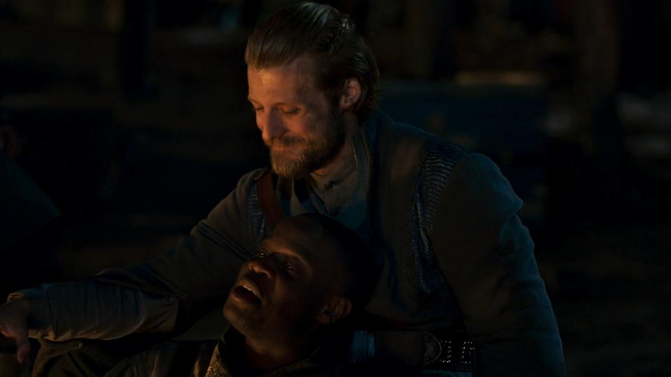 A close-up shot on Maksim (Taylor Napier) and Ihvon (Emmanuel Imani), as they sit around a fire cuddling. Maksim is smiling down at Ihvon, who has his head propped against Maksim’s chest