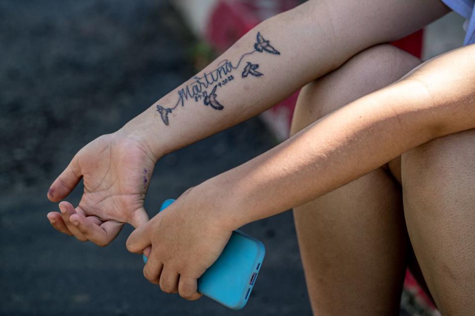 A girl holds her phone and stretches one of her fingers, showing a tattoo of the name Martina and doves on her forearm