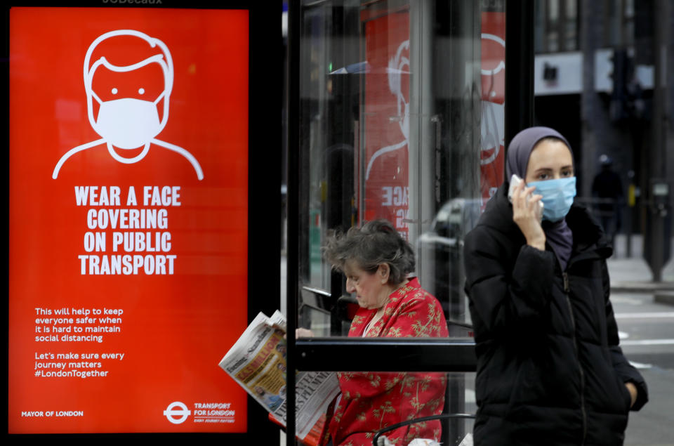 Passengers wait at a bus stop with a sign advising travellers to wear a face covering whilst travelling, in London, Friday, June 5, 2020. It will become compulsory to wear face coverings whilst using public transport in England from Monday June 15. (AP Photo/Kirsty Wigglesworth)