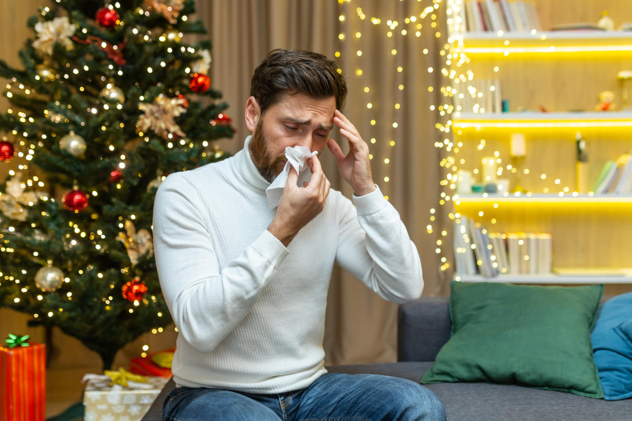 A man wearing a white turtleneck sitting in front of a decorated Christmas tree in a living room, holding a tissue up to his nose and appearing sickly