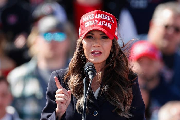 Kristi Noem wearing a "Make America Great Again" hat speaking into microphone, gesturing with one finger raised