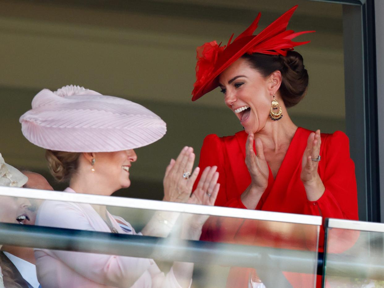 Sophie, the Duchess of Edinburgh, and Kate Middleton smiling and clapping their hands while looking out across a glass balcony.