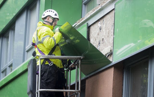 A worker in a hi-vis jacket and a white hard hat removes a green panel from a building