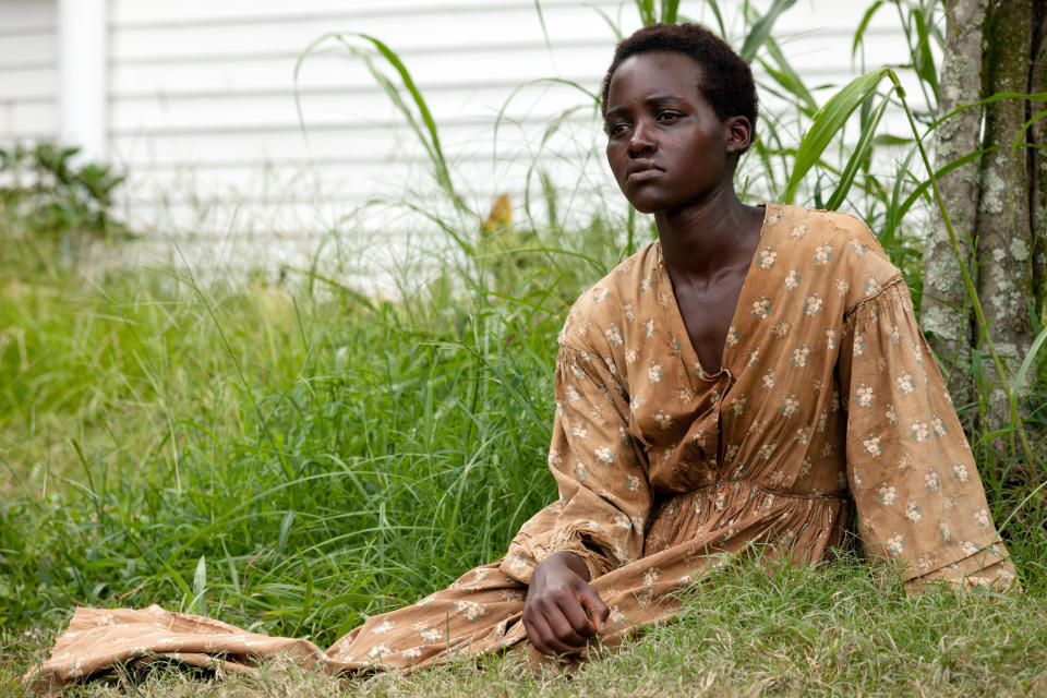 Lupita sitting on grass, leaning on a tree, wearing a long, patterned dress in scene from 12 Years a Slave