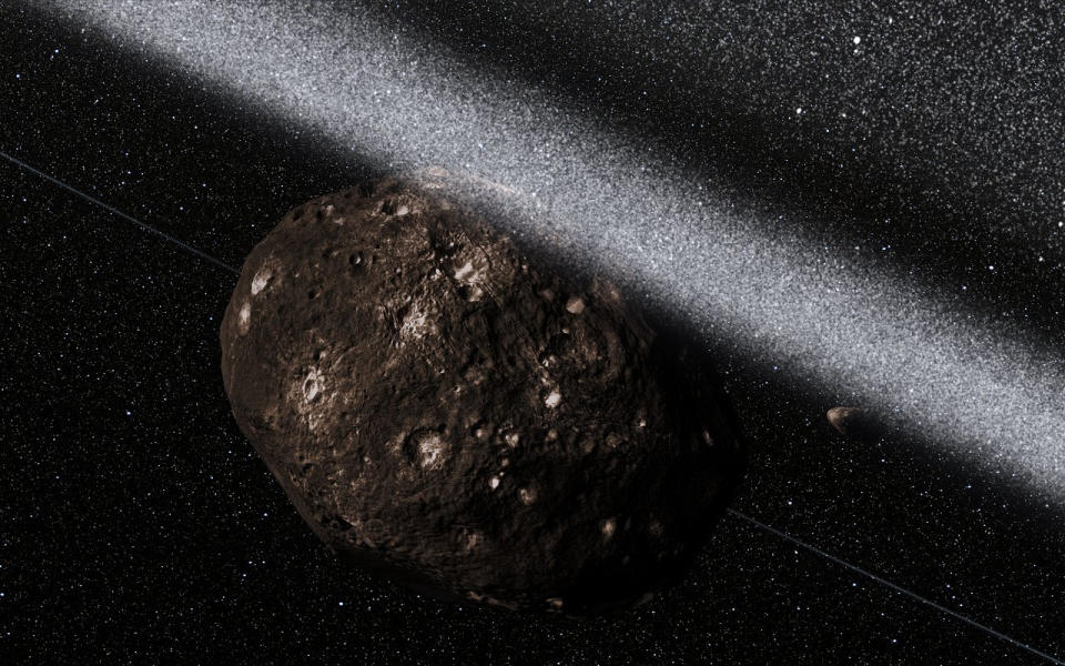 An uncharged but crater-riddled space rock lies far left of center.  The black background of space is full of faint stars, and dusty white rushes form a thick, almost solid line that cuts the image diagonally from top left to center right.