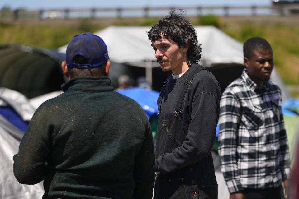 Volunteer Ian Greer talks with an asylum-seeker at an encampment set up next to an unused motel owned by the county, Wednesday, June 5, 2024, in Kent, Washington. The group of about 240 asylum-seekers is asking to use the motel as temporary housing while they look for jobs and longer-term accommodations. (AP Photo/Lindsey Wasson)
