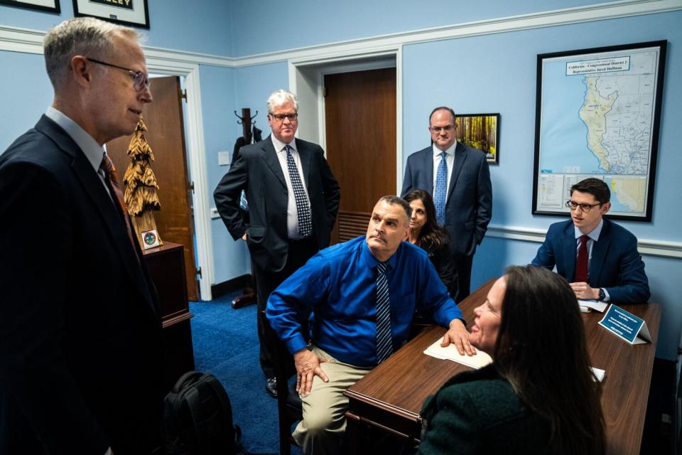 People, dressed in suits, stand or sit around a desk.