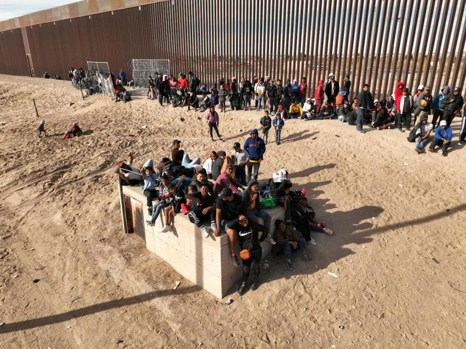 A large group of people are seen sitting and standing along a tall brown fence in an empty area of brown dirt.