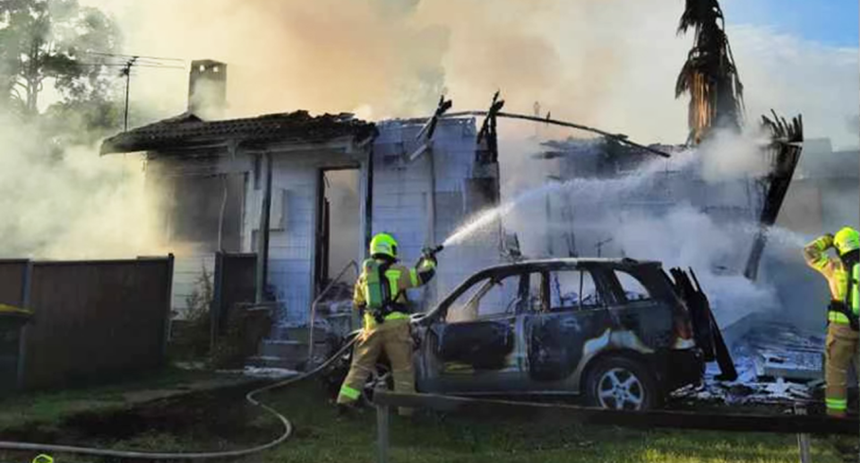 Firefighters direct water onto the smoking building from a hose, with a car burnt and the house ruined. 