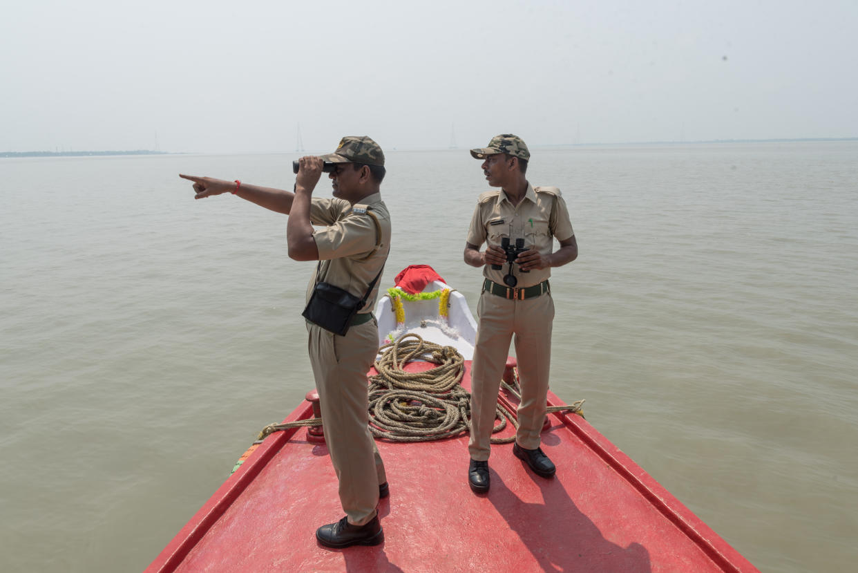 Two officers in peaked caps, both holding binoculars, stand on a bright red police boat in an expanse of water, as one points into the distance.