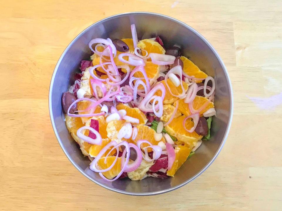 Bird's-eye-view of a metal bowl filled with salad ingredients, including arugula, orange wedges, sliced shallots, and olives