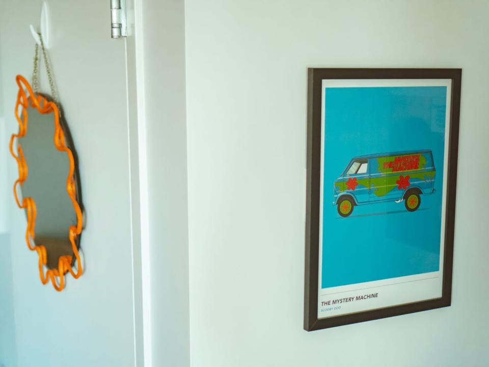 A mirror with an orange squiggle-style frame and a print of the mystery machine from "Scooby-Doo" hang on walls