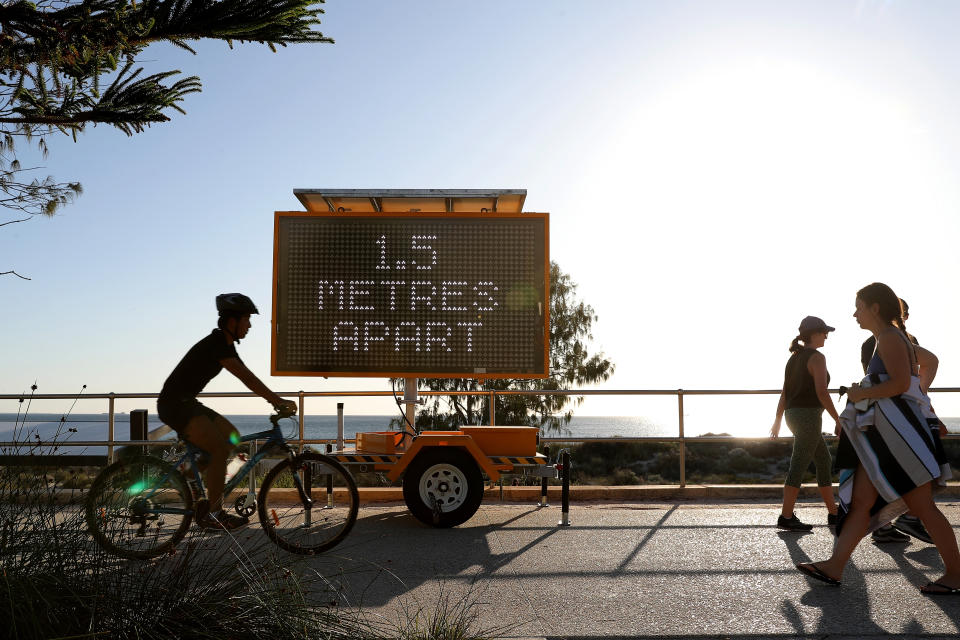 A cyclist and three women walk past a sign reading, '1.5 Metres Apart', at Perth's Scarborough Beach.