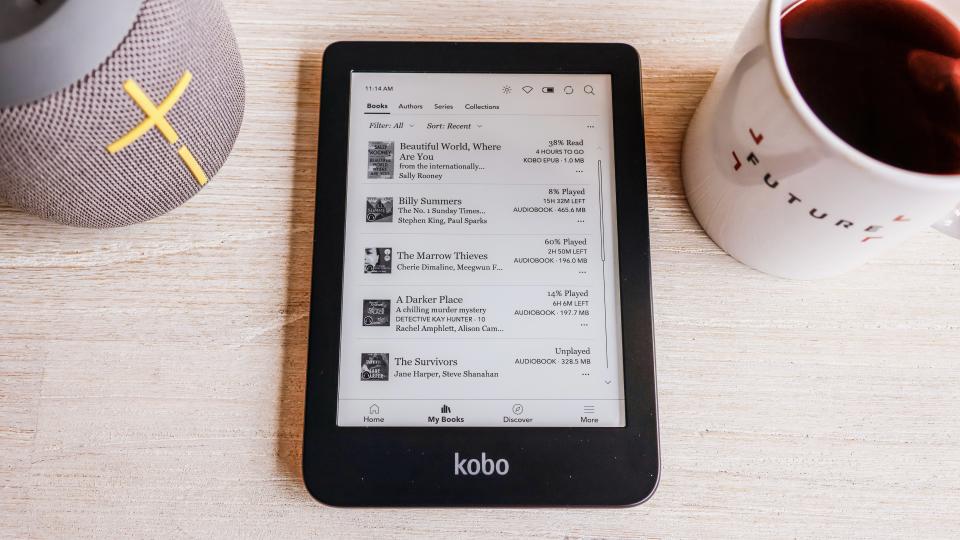 The Kobo Clara 2E laying flat on a table next to a speaker and coffee mug, showing the collection of books loaded onto the device.