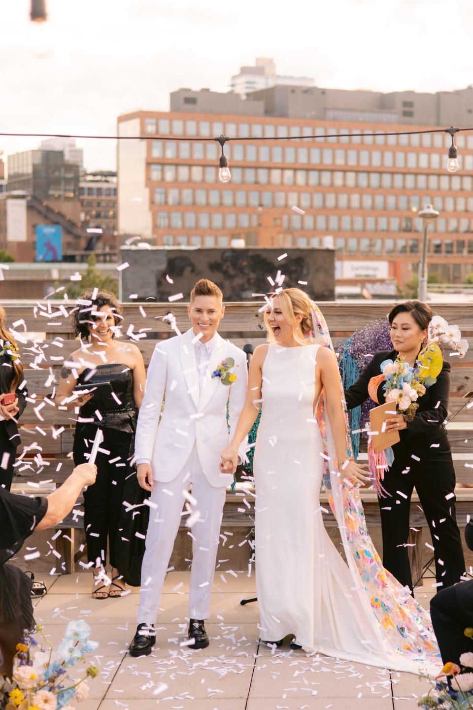 A bride in a white suit and a bride in a white dress and rainbow veil hold hands as confetti explodes around them during their wedding ceremony.