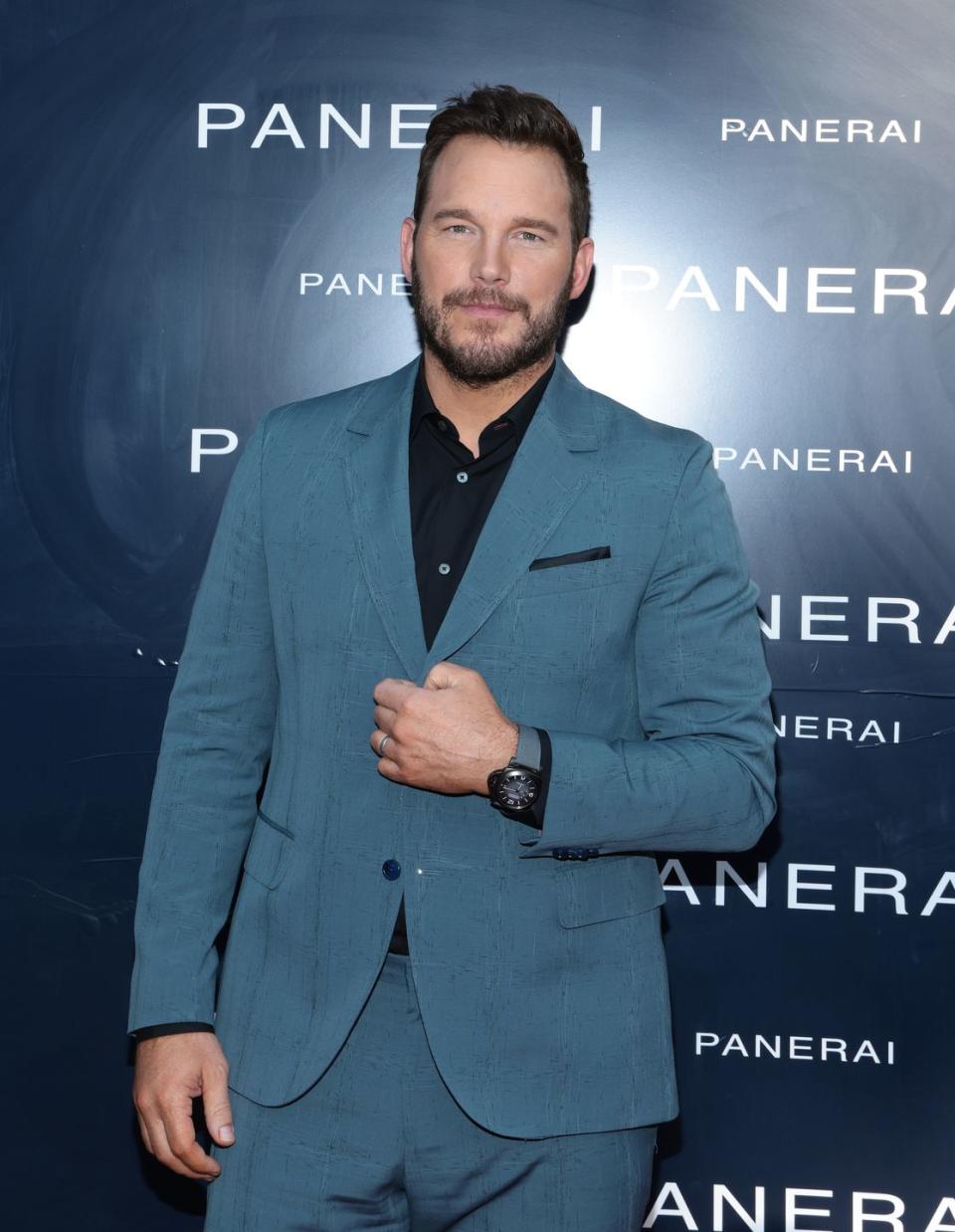 Chris Pratt, a man stands there and looks into the camera with a neutral expression, he has short dark brown hair and a beard, he is wearing a black shirt with a blue jacket