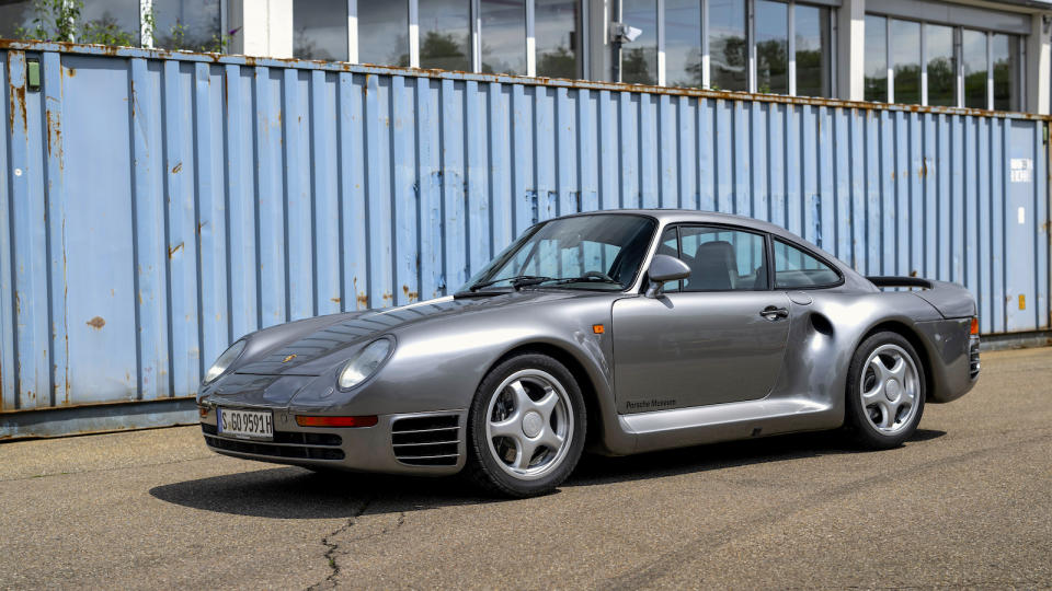 A dark silver Porsche 959 is parked in front of a shipping container.