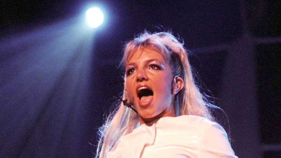 britney spears sings into a headset microphone while lights shine on her, she wears a white pleather jacket and matching pants with pink accents