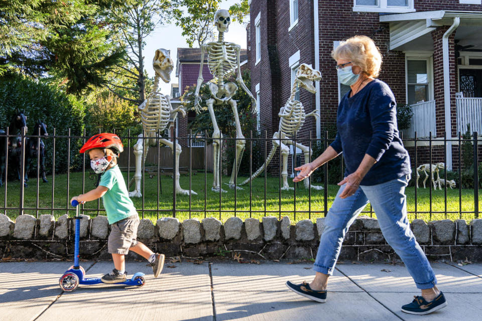 Sam McNamara, 3, of Washington, left, followed by his grandmother, scoots by a yard decorated with giant skeletons, both human and dinosaur, Wednesday, Oct. 14, 2020, in advance celebration of Halloween in Washington. (AP Photo/Jacquelyn Martin)