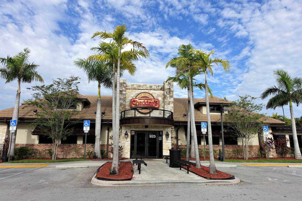 Front exterior of Cheddar's Scratch Kitchen restaurant in Wellington, Florida, front doors, garden, and handicapped parking spots lined by palm trees with an expansive white clouded blue sky in the background
