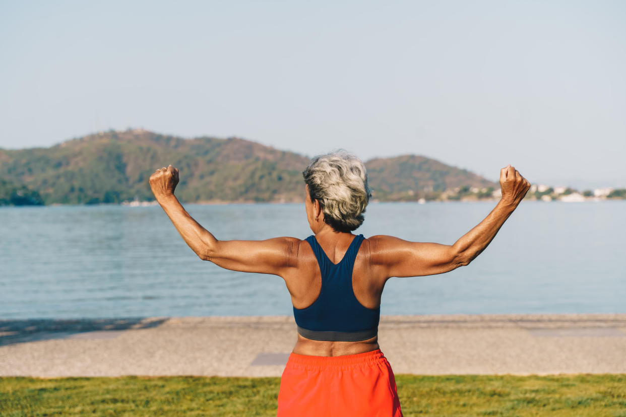 Senior gray-haired woman doing exercises in public park, sunny summer morning. Rear view.