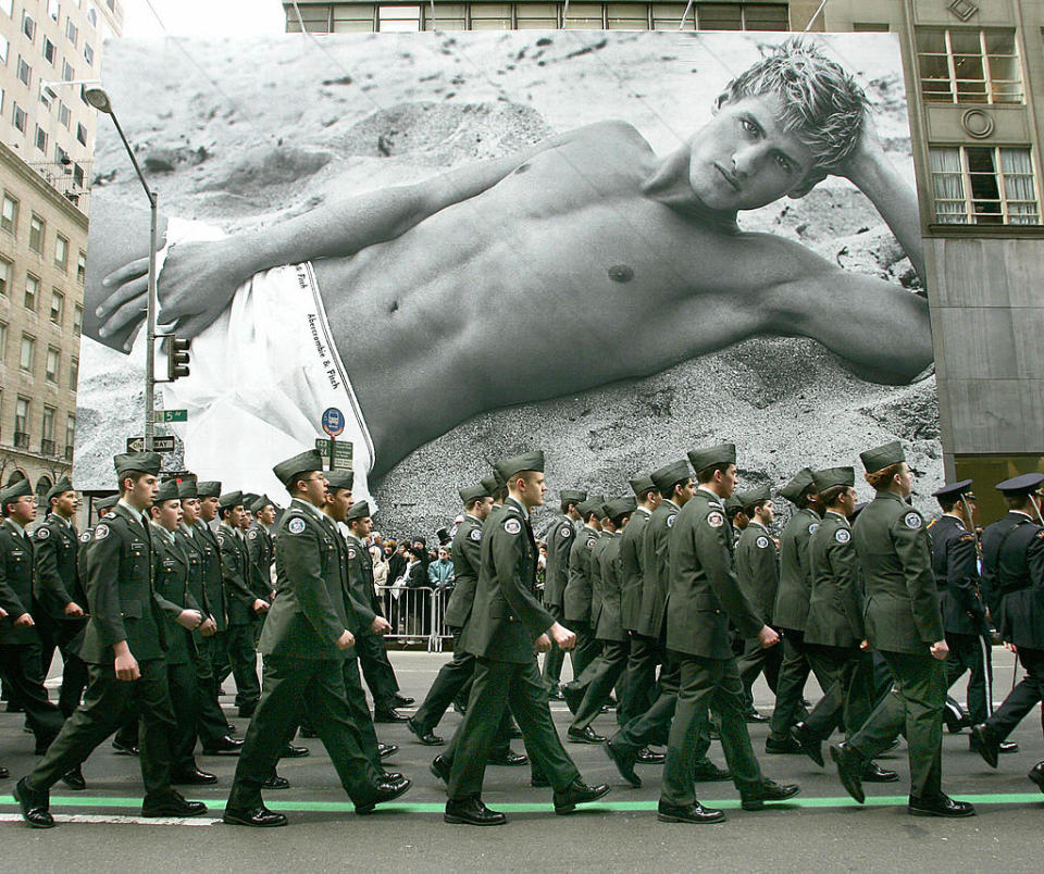 A large group of uniformed military personnel marches in a city. Behind them is a large billboard featuring a black-and-white image of a shirtless man on a beach