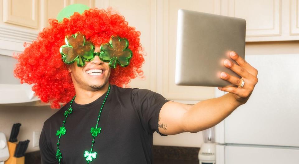 man wearing red wig and st patricks day necklace taking selfie on tablet