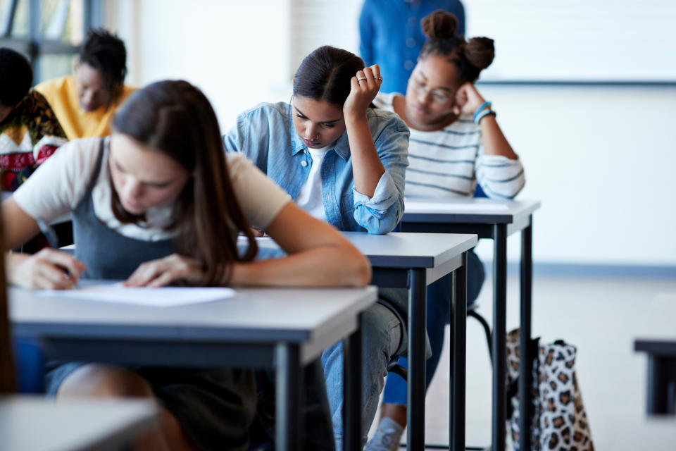 Students sitting at desks; one writing intently, another appears to be thinking hard, with more in the background