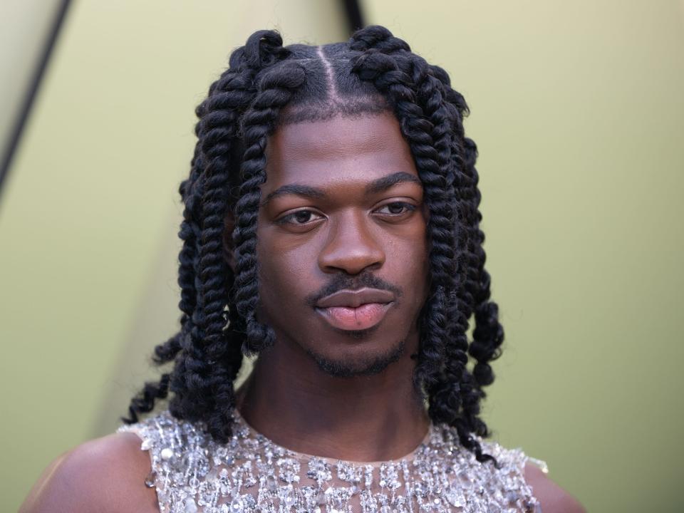 Lil Nas X standing in front of a light green background while wearing a sleeveless top.